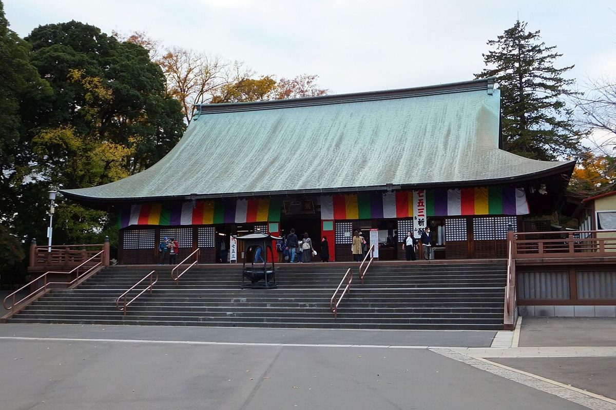 緑青色の大屋根の本堂と石段。軒下に五色の幕と「七五三」の幟が下がり、左奥の木々が黄色く色づいている