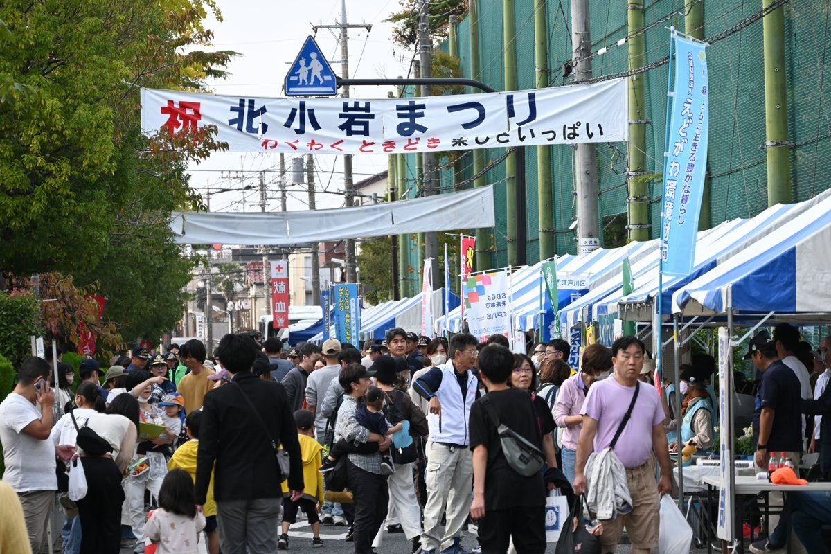 「祝 北小岩まつり」の横断幕の下、中学校のフェンス沿いに白と青のテントが並び、家族連れが歩く道路