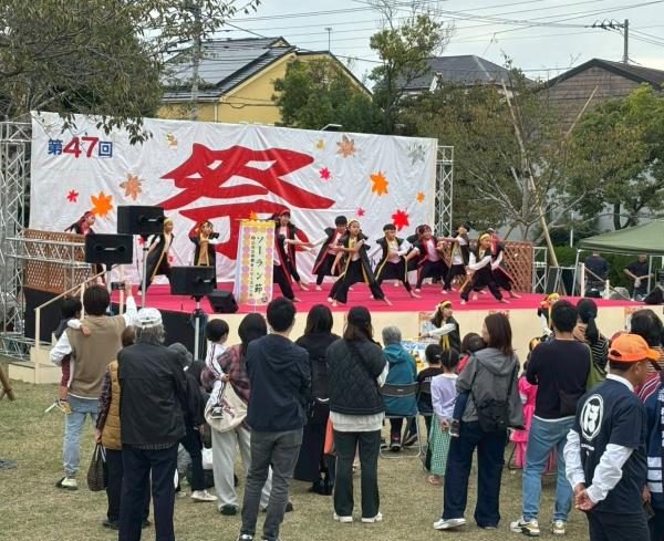 「第47回」と「祭」の幕が張られた屋外ステージで、黒い衣装の子どもたちが踊り、芝生の上で観客が立って見ている