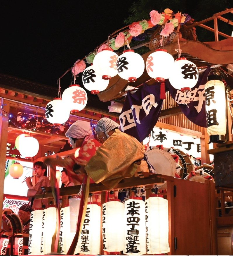 提灯をともした北本四丁目囃子連の囃子山車と、山車の上で面をつけて踊る人。奥に原組の山車
