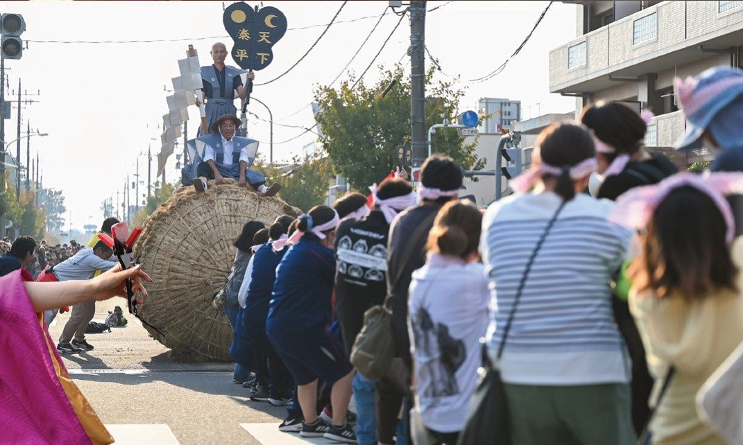 北本駅西口の交差点で、大俵の上に乗った会津坂下町の人と、綱を引く鉢巻き姿の参加者たち