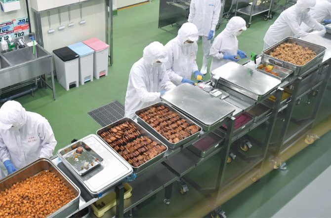 白い作業着の人たちがライン沿いに並び、バットに入ったおかずを弁当に詰めている