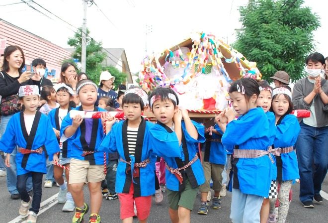 青い法被を着た子どもたちが、色とりどりの飾りを付けた手作りのみこしを担いで通りを進む様子