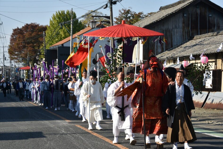 赤い傘を差しかけられた白装束の人や、赤い装束で面をつけた人、のぼり旗を持つ人たちが、商店の並ぶ通りを列になって歩く昼の写真
