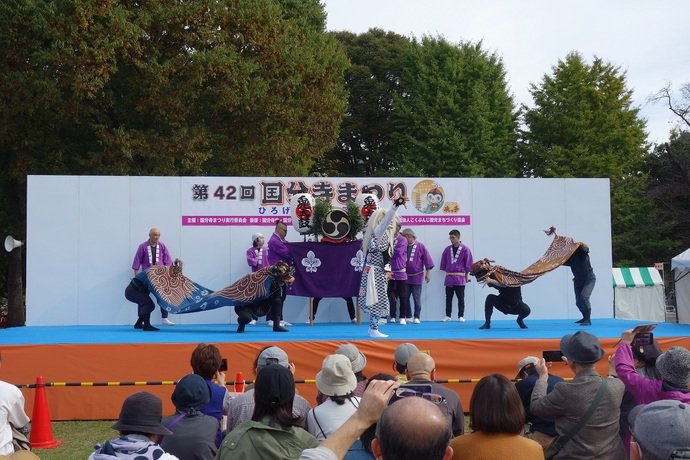 第42回国分寺まつりの看板があるステージで、紫の法被の人たちの前で獅子舞が演じられ、客席から観客が撮影している