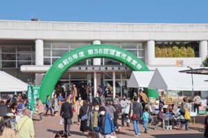 鴻巣市立総合体育館の入口に「令和5年度 第36回鴻巣市産業祭」と書かれた緑のアーチがあり、白いテントの前を多くの来場者が歩いている写真
