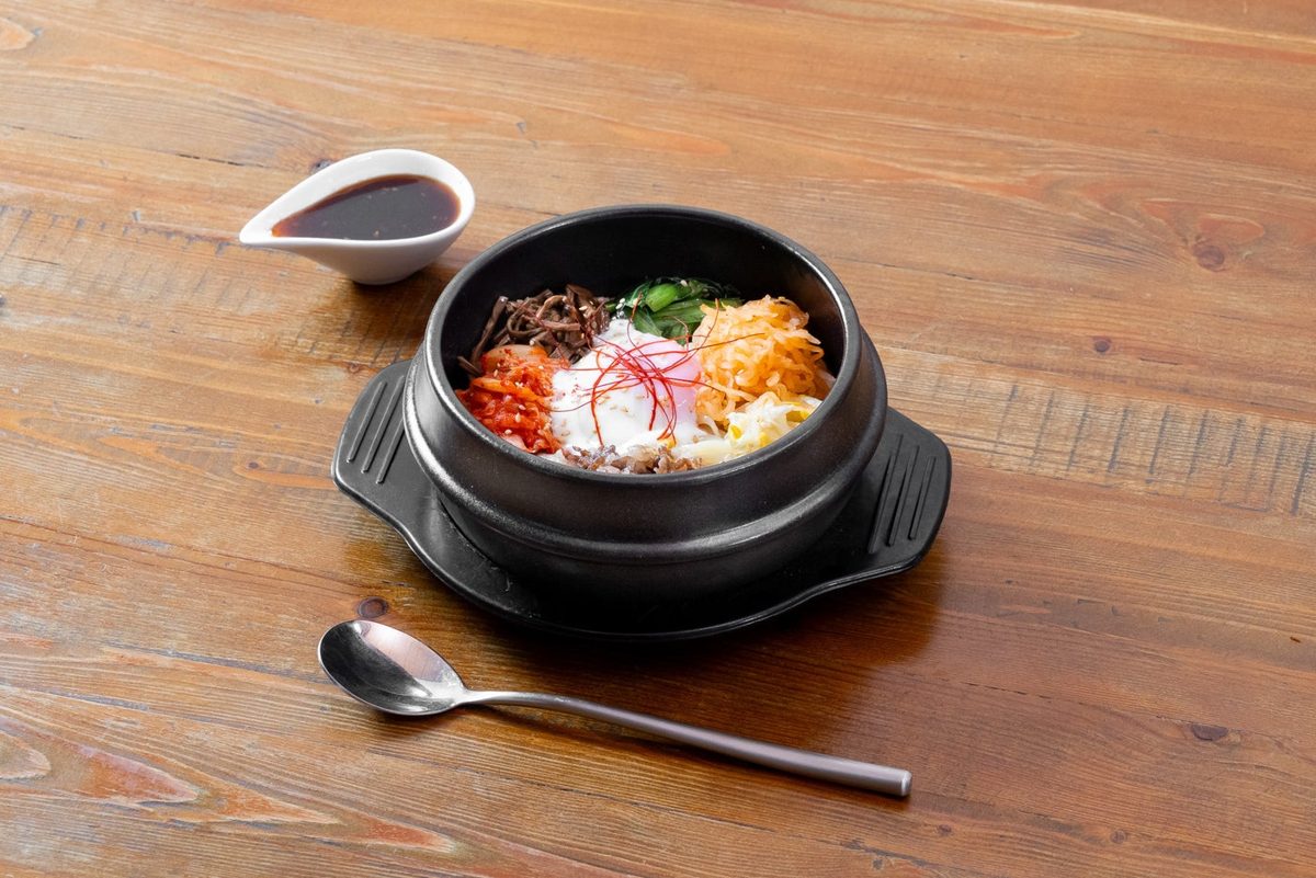 黒い石鍋に盛られたビビンバ。ナムル、キムチ、赤い野菜と卵がのり、横にスプーンとたれの入った器