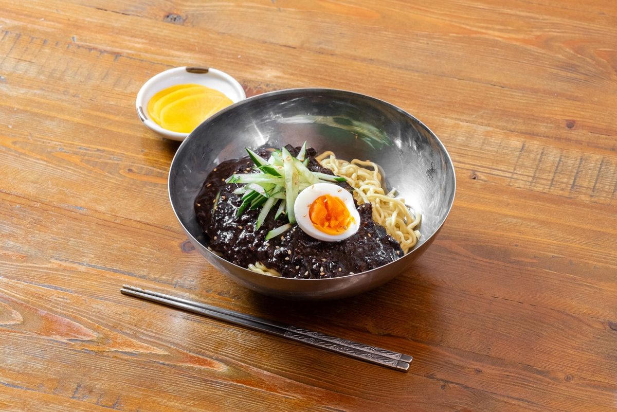 金属の器に盛られた黒い味噌ソースの麺。上に千切りのきゅうりと半分に切ったゆで卵、手前に箸、奥に黄色いたくあん