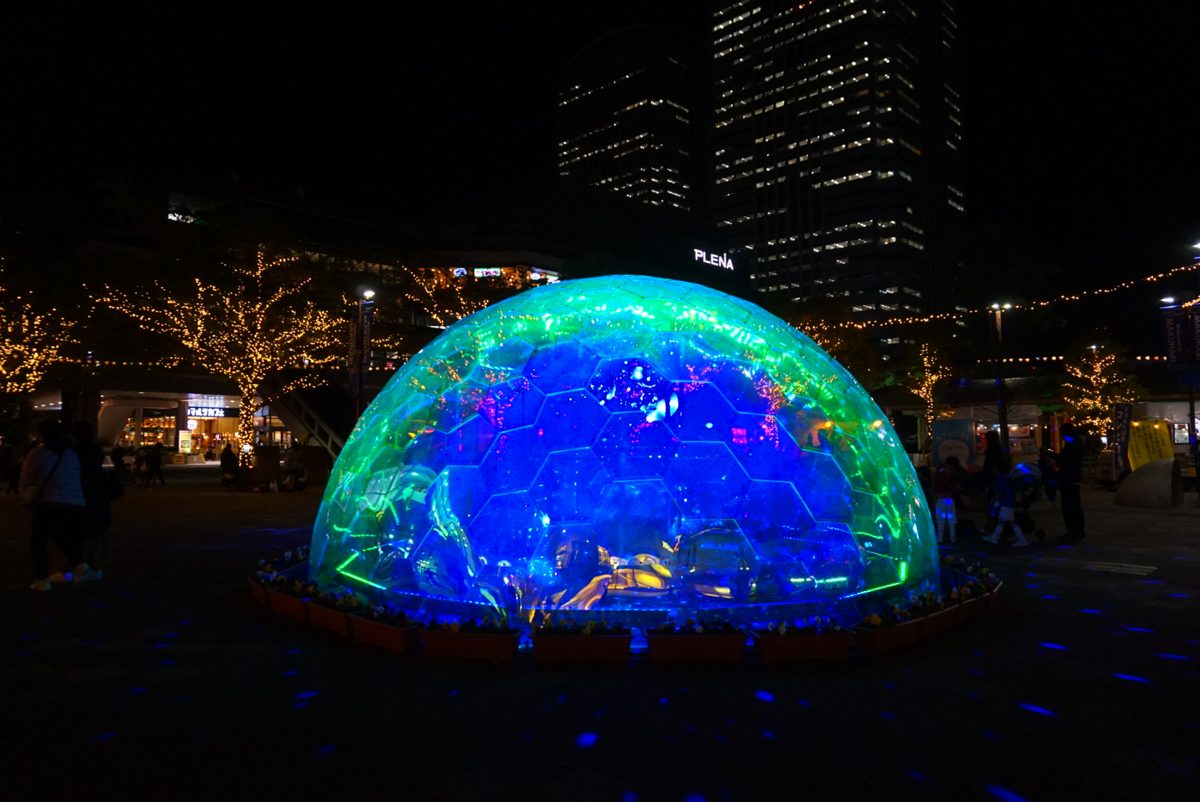 夜の海浜幕張駅南口広場で青や緑に光るレインボードームと、奥の電飾をつけた並木、高層ビル