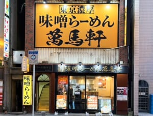 黄色い大きな看板に「東京濃厚 味噌らーめん 萬馬軒」と書かれた店舗の正面。1階の入口の左に2階へ上がる階段がある
