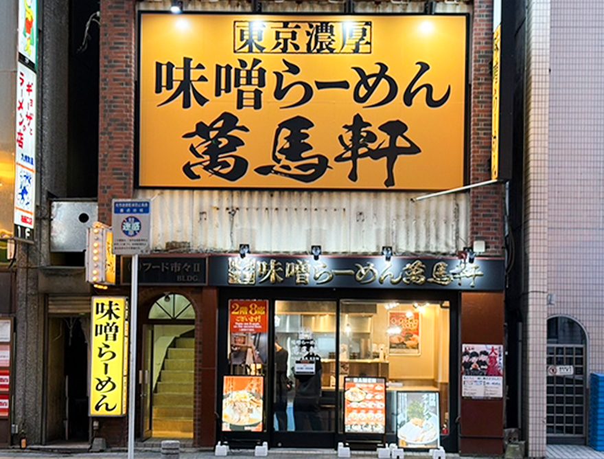 黄色い大きな看板に「東京濃厚 味噌らーめん 萬馬軒」と書かれた店舗の正面。1階の入口の左に2階へ上がる階段がある