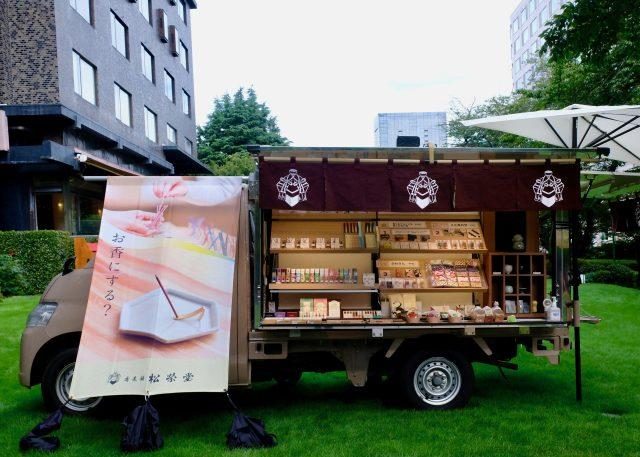 芝生の上に止まった軽トラックの荷台が棚になり、お香の箱が並んでいる。松栄堂の紋の入った暖簾と「お香にする？」ののぼり