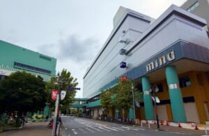 ミーナ津田沼の外観。灰色の外壁に「mina」のロゴと緑色の柱があり、ユニクロとGUの看板が掲げられた建物の前を道路が通っている