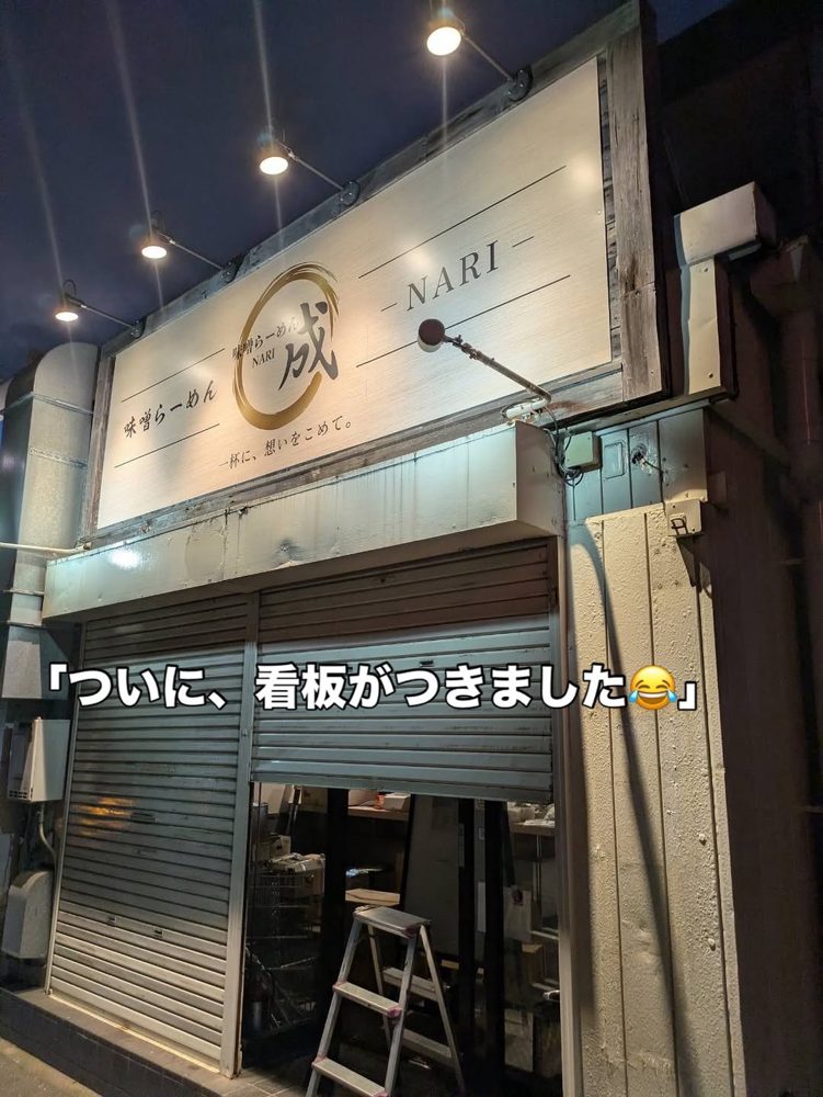2階建ての建物の1階に「味噌らーめん 成 -NARI-」の白い看板が付き、シャッターが半分下りた開店前の店先。写真に「ついに、看板がつきました」の文字