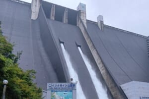 曇り空の下、宮ヶ瀬ダムの堤体から2本の水が放流され、手前にインクライン山麓駅の看板が立っている写真