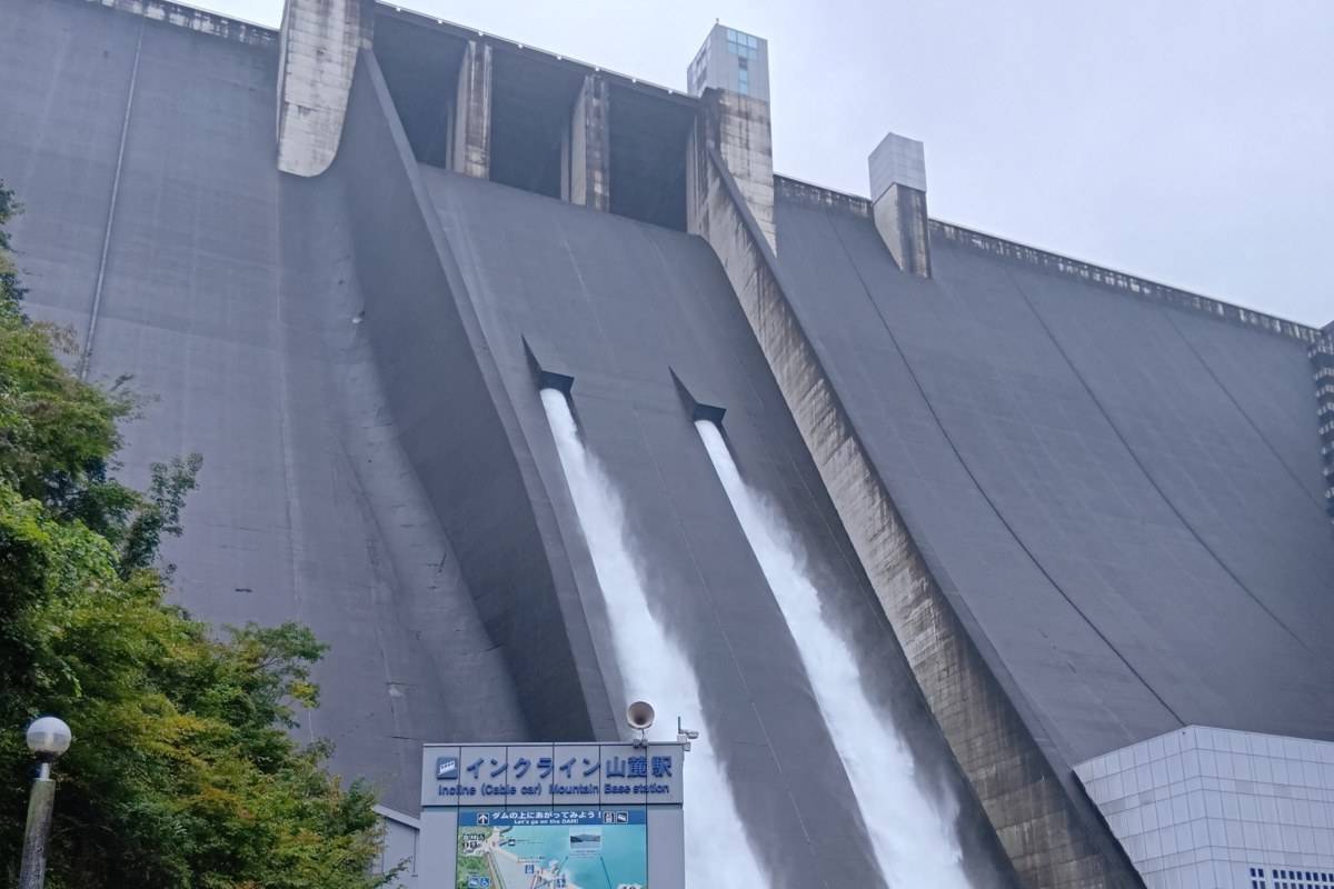 曇り空の下、宮ヶ瀬ダムの堤体から2本の水が放流され、手前にインクライン山麓駅の看板が立っている写真