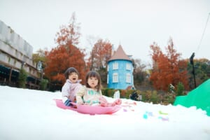 紅葉した木々と青いムーミン屋敷を背に、雪の上でピンクのそりに乗って笑う2人の子どもの写真