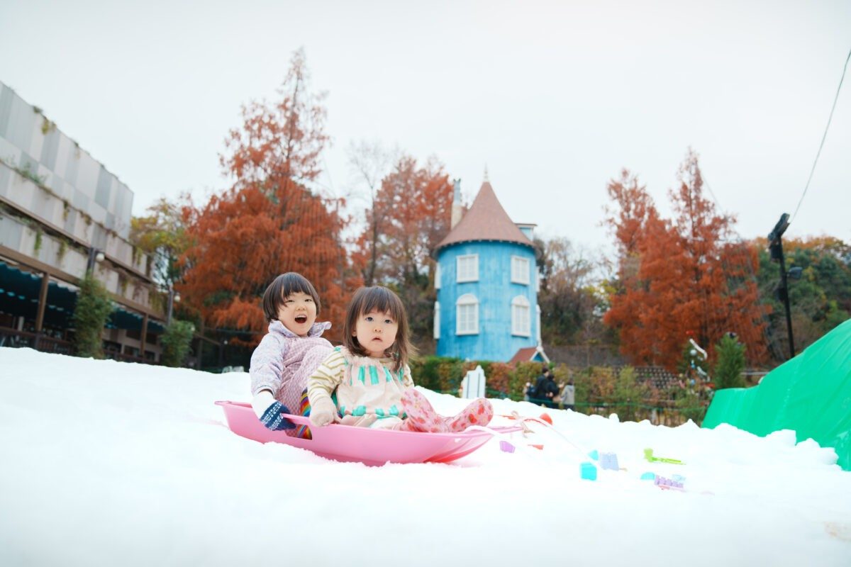 紅葉した木々と青いムーミン屋敷を背に、雪の上でピンクのそりに乗って笑う2人の子どもの写真