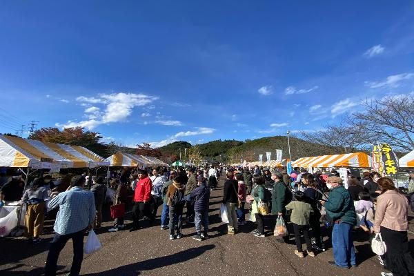 青空の下、黄色と白のテントが並ぶ会場の通路を多くの来場者が歩いている2023年の毛呂山町産業まつりの写真