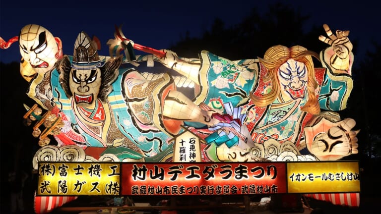 夜の闇に照らし出された、武者の顔や鬼の顔が描かれた大きな山車。下に「村山デエダラまつり」の看板が付いている