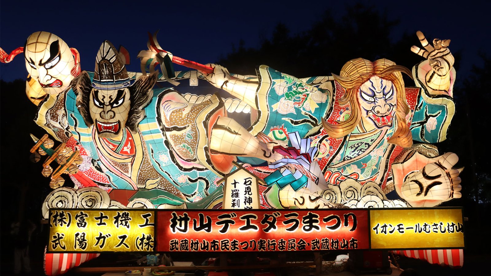 夜の闇に照らし出された、武者の顔や鬼の顔が描かれた大きな山車。下に「村山デエダラまつり」の看板が付いている