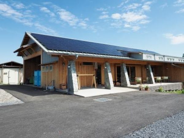 青空の下に建つ、太陽光パネルを載せた木造の平屋の建物と、手前の舗装された敷地