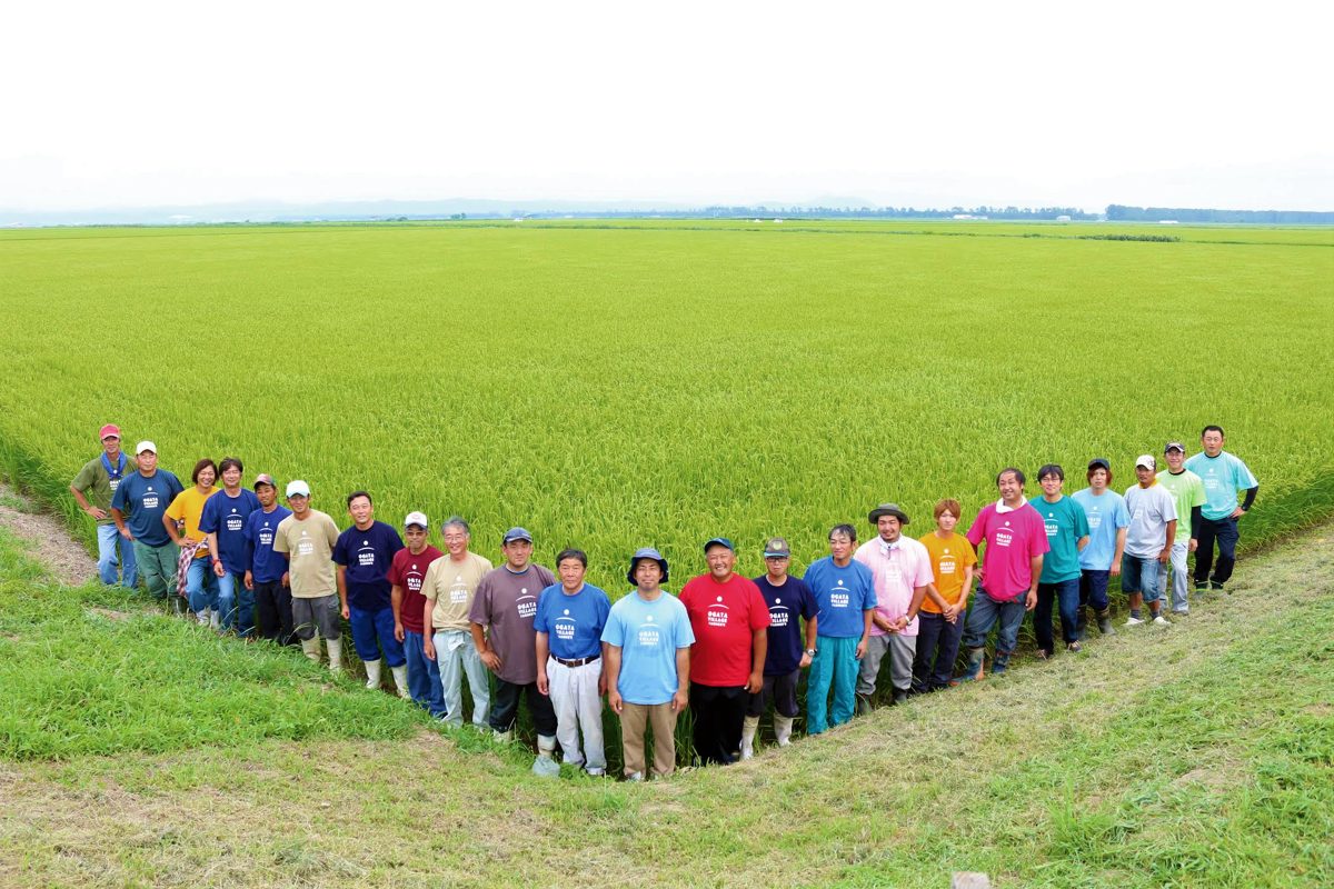 一面の緑の田んぼの前に、色とりどりのTシャツの生産者約20人が並ぶ写真