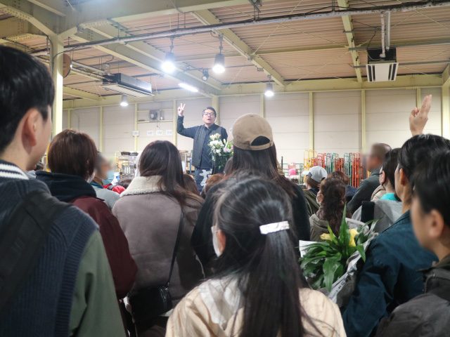 花束を持って手を挙げるせり人の前に来場者が集まり、手を挙げて花を競り落とそうとしている