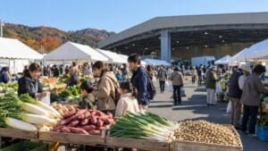 卸売市場の前に白いテントが並び、大根やさつまいも、ねぎ、落花生を家族連れが選んでいるイメージ画像（AI生成）