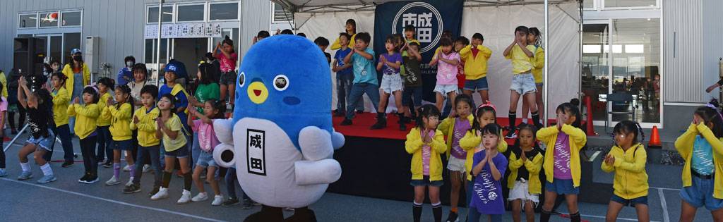 成田市場の建物前の特設ステージで、うなりくんと黄色い服の子どもたちが踊っている