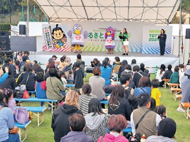 屋外テントのステージで、ねんりんピックのキャラクターの着ぐるみと司会者が立ち、ベンチの観客が見ている写真。ステージ右の背面に「NENRIN TOTTORI 2024」の表示