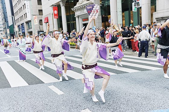 日本橋・京橋まつりの大江戸活粋パレード。紫と白の衣装の阿波おどりの踊り手が中央通りの横断歩道を進み、沿道に見物客が並んでいる