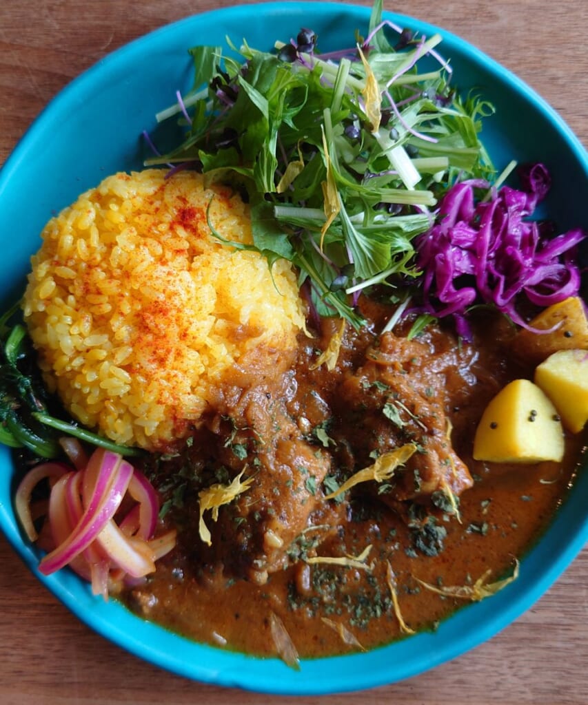 青い皿に盛られたカレーと黄色いライス、水菜と紫キャベツのサラダ、ピクルスを添えたプレート料理