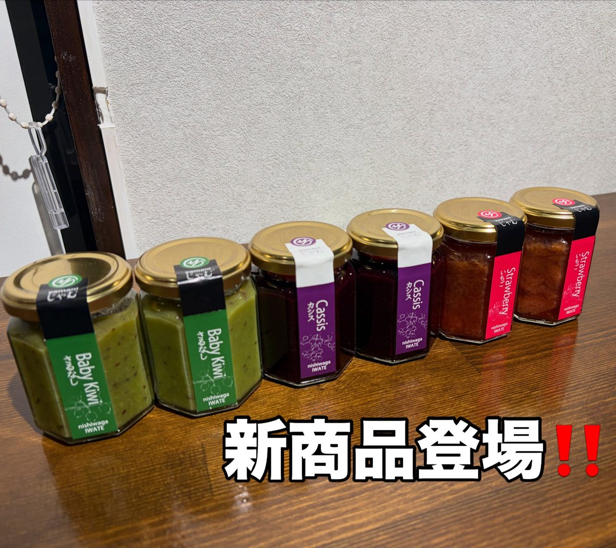 木のテーブルに並んだ緑・紫・赤のラベルのジャムの小瓶6本と「新商品登場!!」の文字