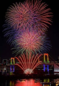 夜空に大きく開いた色とりどりの花火と、レインボーブリッジの手前で扇状に上がる赤い花火