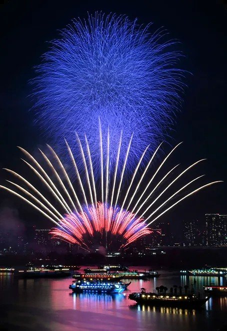 夜空に上がる青い花火と扇状に開く赤や白の花火、手前の海に明かりをともした屋形船が並ぶ様子