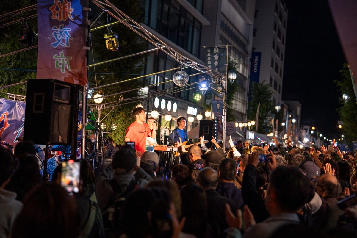 夜の通りに組まれたステージで演奏するバンドと、手を挙げて盛り上がる大勢の観客