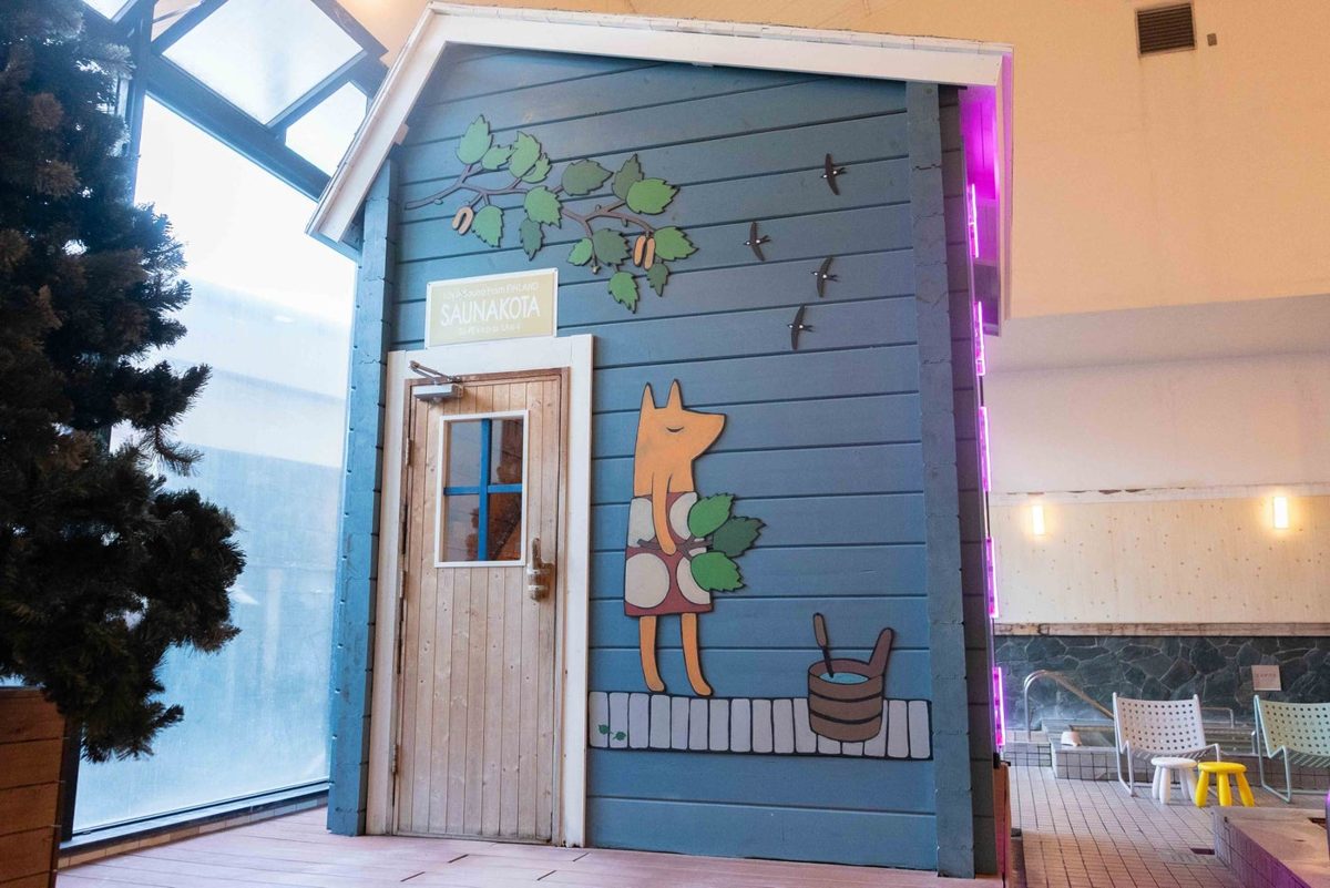 青い板張りのサウナ小屋の外壁に、桶を持ったきつねと葉の枝のアートが描かれ、白い枠の窓と木の扉がある浴室内の様子