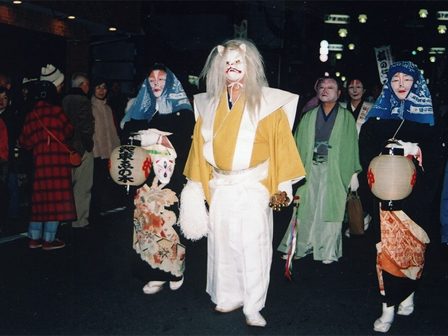 狐の面をつけ、和装で提灯を持って夜の通りを歩く行列の参加者たち