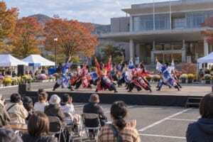 屋外ステージで鳴子を持って踊るよさこいチームのイメージ（AI生成）