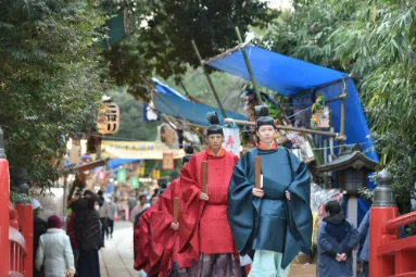 露店の青いテントが並ぶ境内の道を、赤と青の装束を着た神職が歩いている大湯祭の写真