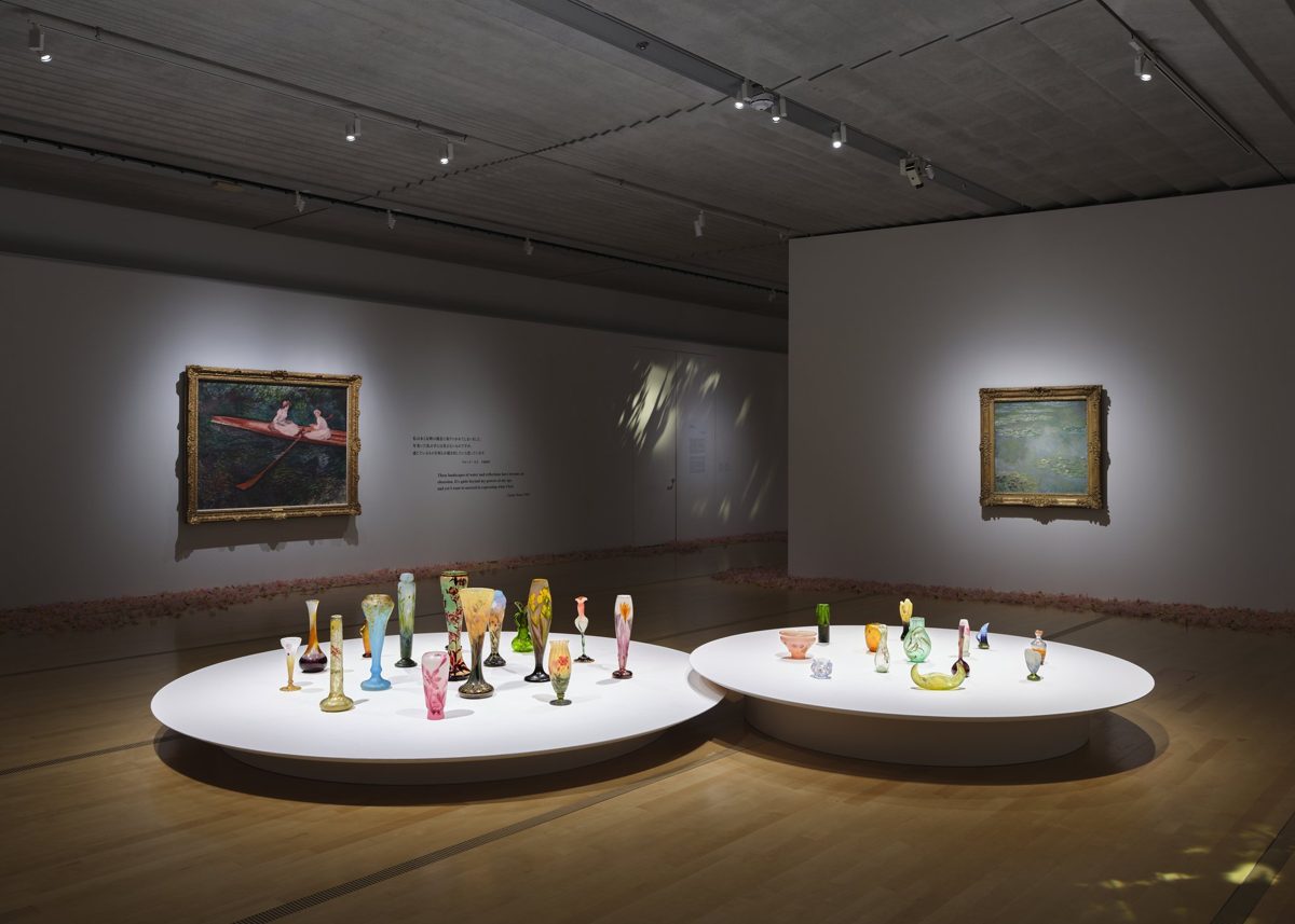 暗い展示室の壁に額装された絵画が掛かり、手前の白い円形の台にガラスの花器が並ぶ