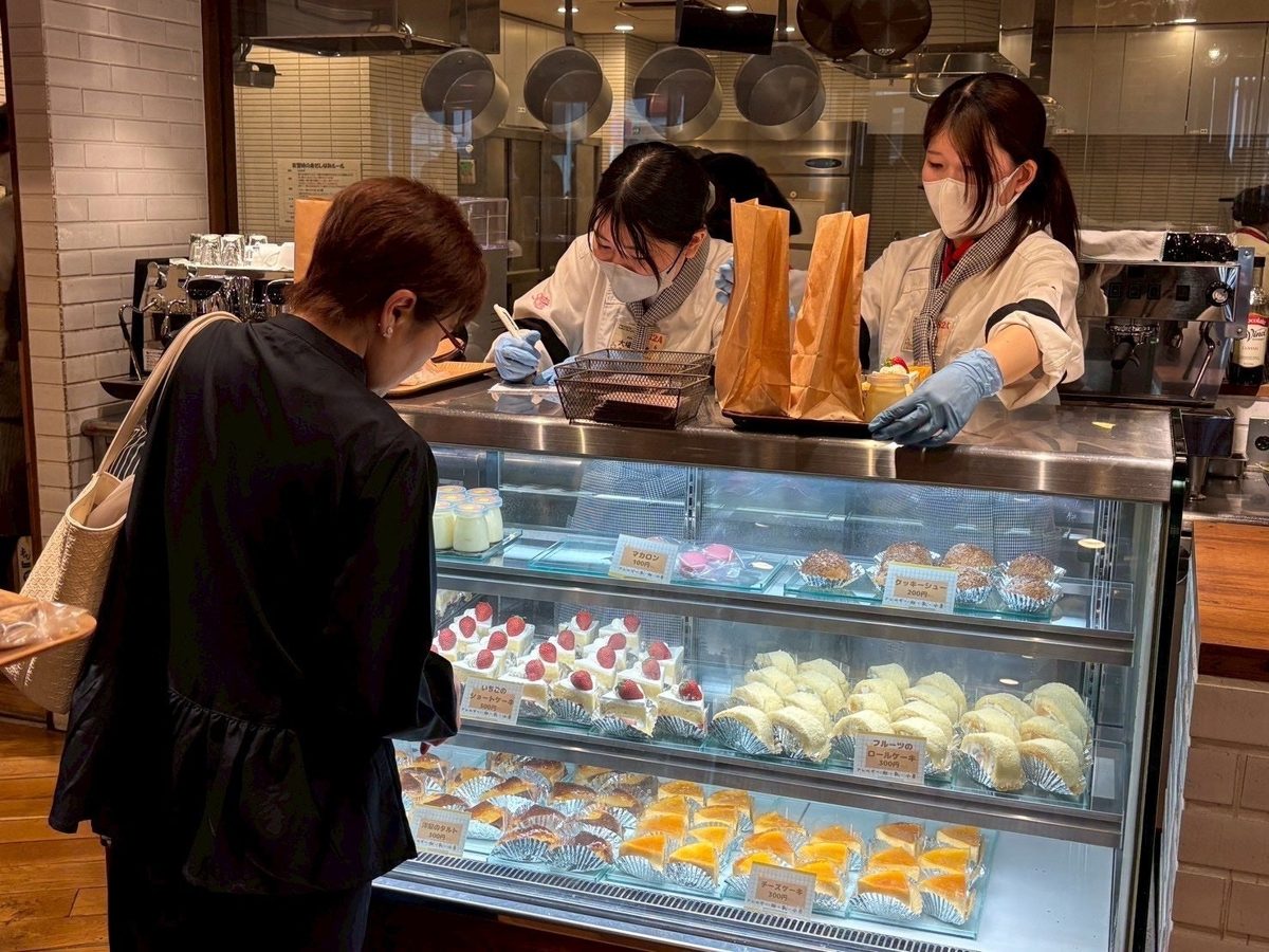 学内店舗のショーケースに並んだケーキを、来店客と白い制服の学生がはさんで選んでいる様子
