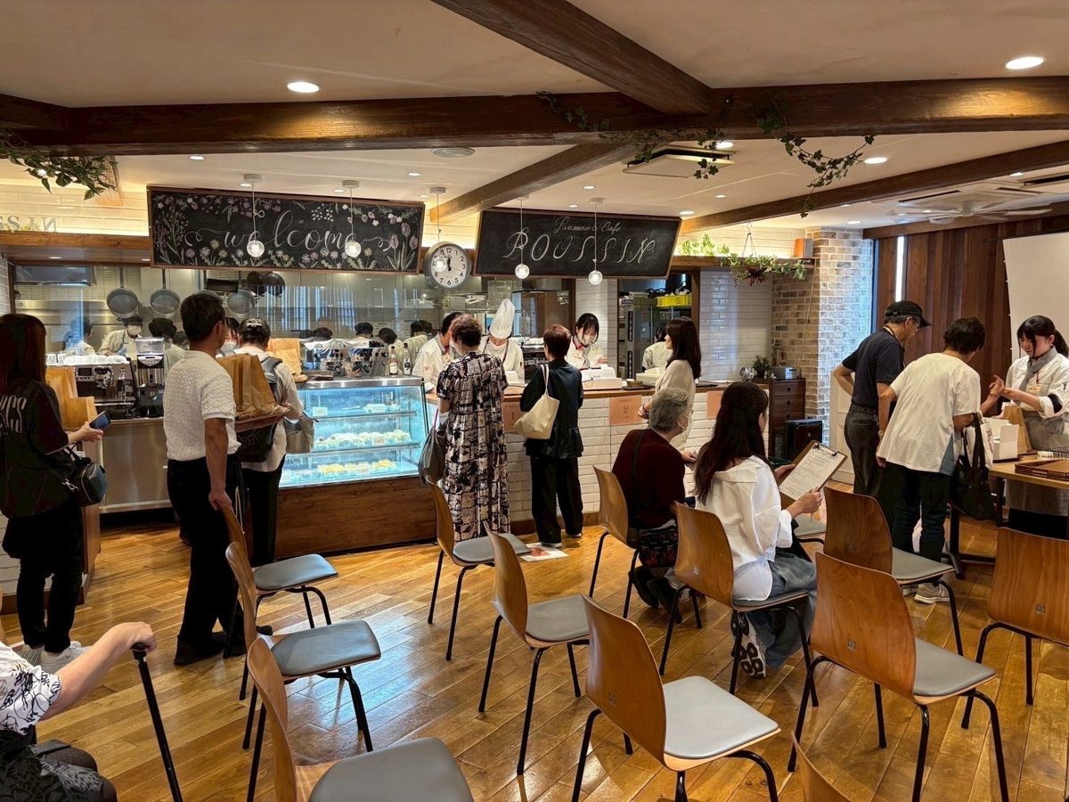 木の床とテーブル席が並ぶ学内カフェの店内で、来店客がショーケースの前に並ぶ様子
