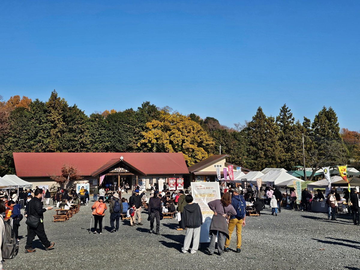 赤い屋根の建物の前の砂利の広場に白いテントが並び、多くの来場者が歩いたりテーブル席に座ったりしている様子