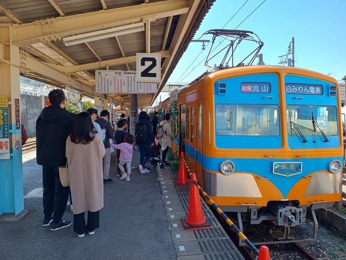 流山駅の2番線ホームに止まるオレンジと水色の流鉄の車両。行き先表示に「白みりん電車」、ホームには買い物に来た人が並ぶ