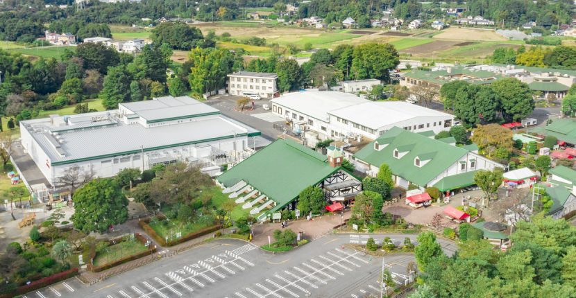 緑の屋根の建物と白い建物、駐車場が並び、周囲に畑と林が広がるサイボクの園内を上空から撮った写真