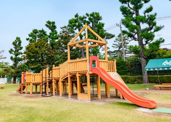 芝生の上に立つ木製の大型アスレチック遊具。オレンジ色の滑り台が付き、後ろに松の木が並ぶ