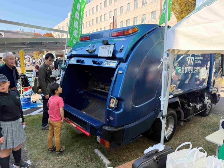 テントの並ぶ広場に止まった青いゴミ収集車(パッカー車)の後ろを、子どもたちがのぞき込んでいる写真