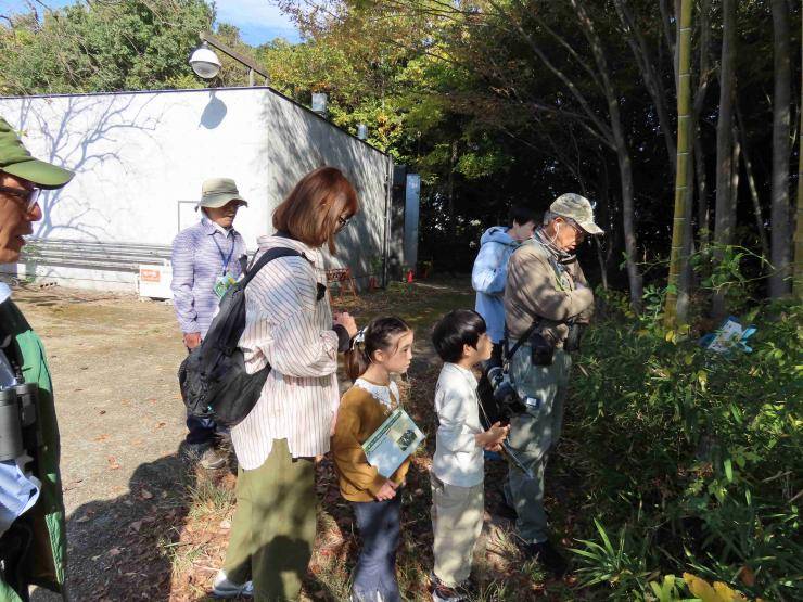 木々に囲まれた生態園で、帽子をかぶったガイドの説明を、双眼鏡を持った大人と子どもたちが聞いている写真
